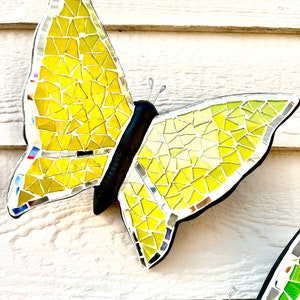 May include: Two mosaic butterflies, one yellow and one green, with black bodies and silver wire antennae. The butterflies are mounted on a white wooden surface.