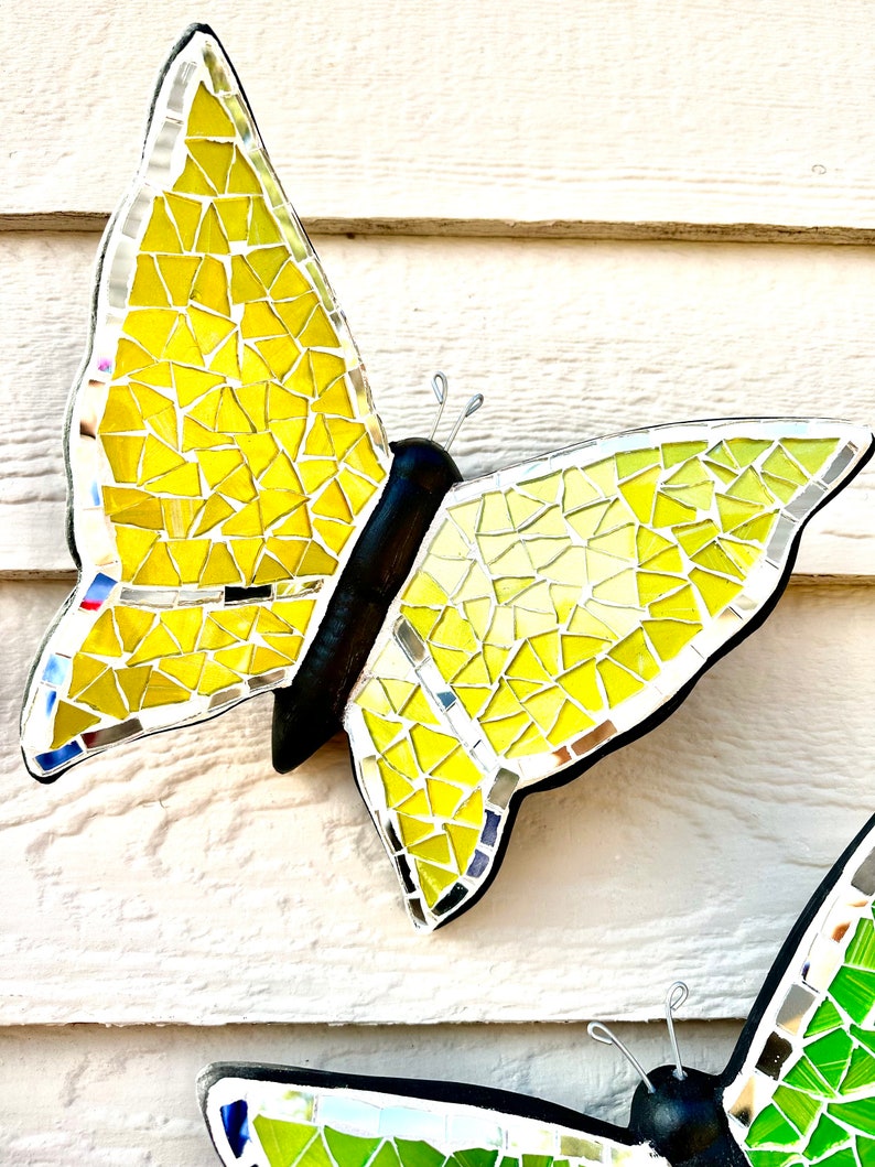 May include: Two mosaic butterflies, one yellow and one green, with black bodies and silver wire antennae. The butterflies are mounted on a white wooden surface.