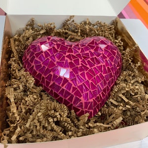 Könnte beinhalten: Eine Mosaik-Herzskulptur in Rosa- und Magentatönen, bestehend aus kleinen, unregelmäßigen Fliesen. Das Herz befindet sich in einer Schachtel, die mit braunem Knitterpapier gefüllt ist. Die Schachtel hat eine rosa Außenseite und eine weiße Innenseite.