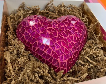 May include: A mosaic heart sculpture in shades of pink and magenta, composed of small, irregular tiles. The heart is nestled in a box filled with brown crinkle paper. The box has a pink exterior and a white interior.