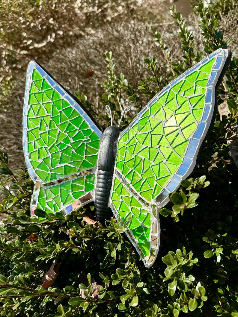 May include: A green mosaic butterfly with a black body. The butterfly is made of small, iridescent glass tiles.