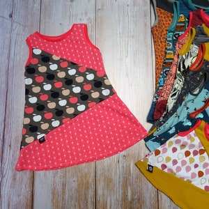 Peut inclure: Une robe sans manches avec un imprimé de pommes rouges, blanches et brunes sur la moitié supérieure et un motif géométrique rose sur la moitié inférieure. La robe est faite d'un tissu doux et extensible et a une coupe ample.