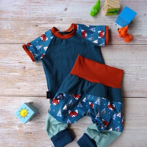 Pode incluir: Um conjunto de bebé de duas peças em verde-azulado, ferrugem e verde claro. A camisa de manga curta tem um corpo verde-azulado com acabamentos em ferrugem e mangas estampadas com raposas vermelhas e triângulos brancos. As calças a condizer têm uma cintura em ferrugem.