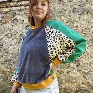 May include: A colour-blocked sweatshirt featuring a dark grey centre panel, leopard print sleeves, and solid green upper sleeve sections. The garment includes a mustard yellow crew neckline, cuffs, and hem, with a small circular patch on the hem.