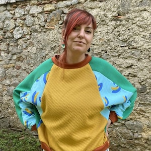 Op de afbeelding: Een sweatshirt met lange mouwen voorzien van een gewatteerd mosterdgeel middenpaneel, lichtblauwe mouwen met een herhalend geel en donkerblauw abstract patroon, effen groene schouderstukken en een donkeroranje geribbelde ronde hals en boorden.