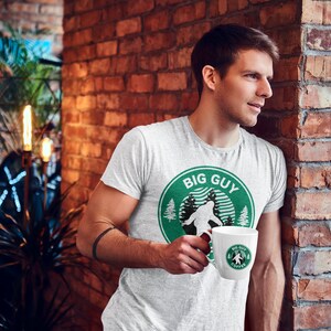 Puede incluir: Una camiseta blanca con un gráfico circular verde que dice "Big Guy Coffee" y presenta una silueta de Bigfoot. La persona sostiene una taza blanca con el mismo gráfico.