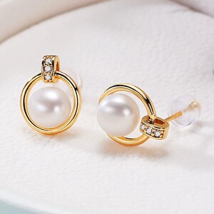 May include: A pair of gold-colored stud earrings featuring a large, round, white pearl suspended within a gold hoop. The top of the hoop is accented with small, clear gemstones. The earrings have a classic and elegant design.