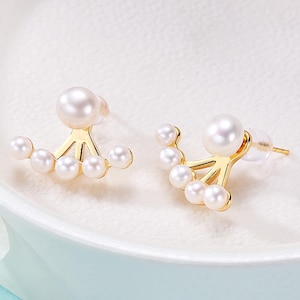 May include: A pair of gold-colored earrings featuring a large pearl on top and a fan-shaped arrangement of smaller pearls below. The earrings are set against a soft, white background, highlighting their elegant design.