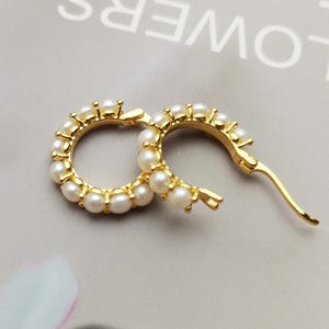 May include: A pair of gold-colored hoop earrings, each adorned with small, round, white pearls. The earrings are set against a soft, neutral background, highlighting their elegant design. The clasp is visible on one earring.