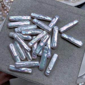 Puede incluir: Un conjunto de perlas rectangulares iridiscentes. Las perlas tienen un brillo nacarado y lustroso con toques de rosa, azul y verde. Están dispersas sobre una superficie gris, lo que sugiere un entorno de fabricación de joyas. Las perlas son de diferentes longitudes.