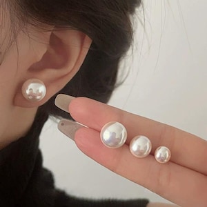 Może przedstawiać: Kolekcja białych kolczyków z pereł w różnych rozmiarach. Jeden duży kolczyk z perłą jest noszony na uchu, a trzy inne kolczyki z perłami o malejącym rozmiarze są trzymane w dłoni. Perły mają połyskującą, opalizującą poświatę.