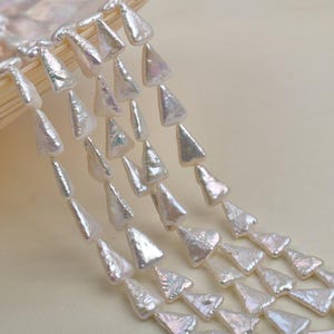 Puede incluir: Primer plano de varias hebras de perlas blancas de forma triangular. Las perlas tienen un brillo iridiscente y se muestran sobre un fondo neutro. Las perlas están dispuestas en un patrón en cascada, ideal para el diseño de joyas.