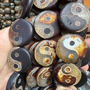 Puede incluir: Una colección de cuentas de piedra marrón oscuro y bronceado, cada una con un símbolo de Yin y Yang. Las cuentas tienen forma circular con una superficie lisa y pulida. Los símbolos de Yin y Yang son de un color claro contrastante.