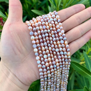 Op de afbeelding: Strengen glanzende, veelkleurige parels. De parels vertonen een reeks kleuren, waaronder wit, roze en goud. De parels worden in een hand gehouden tegen een achtergrond van groen gebladerte. De parels zijn een klassiek sieraad.