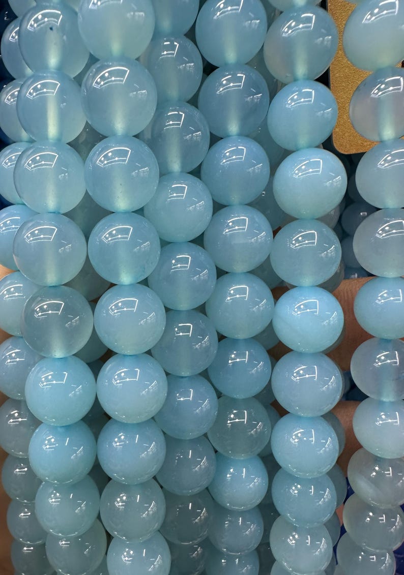 May include: A close-up of a strand of light blue, round beads. The beads are smooth and translucent, with a slight sheen. The beads are strung together, creating a continuous line of color and texture. The beads are a soft, pastel blue.