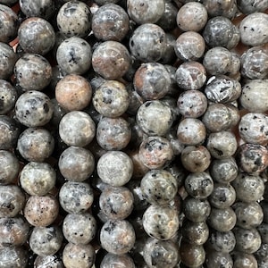 May include: Close-up of a collection of round, speckled beads. The beads are primarily gray with brown and black flecks, creating a natural, textured appearance. These beads are likely used for jewelry making.