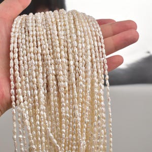 Può includere: Un gruppo di perle d'acqua dolce bianche crema, di forma ovale. Le perle sono infilate insieme, formando più fili. Le perle hanno una lucentezza naturale e dimensioni variabili. Lo sfondo è sfocato, enfatizzando le perle.