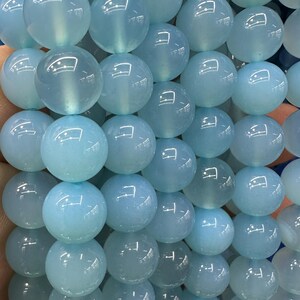 May include: Close-up of a strand of light blue, round gemstone beads. The beads are smooth and translucent, with a subtle sheen. The beads are strung together, creating a full, textured appearance. The beads are approximately 10mm in diameter.