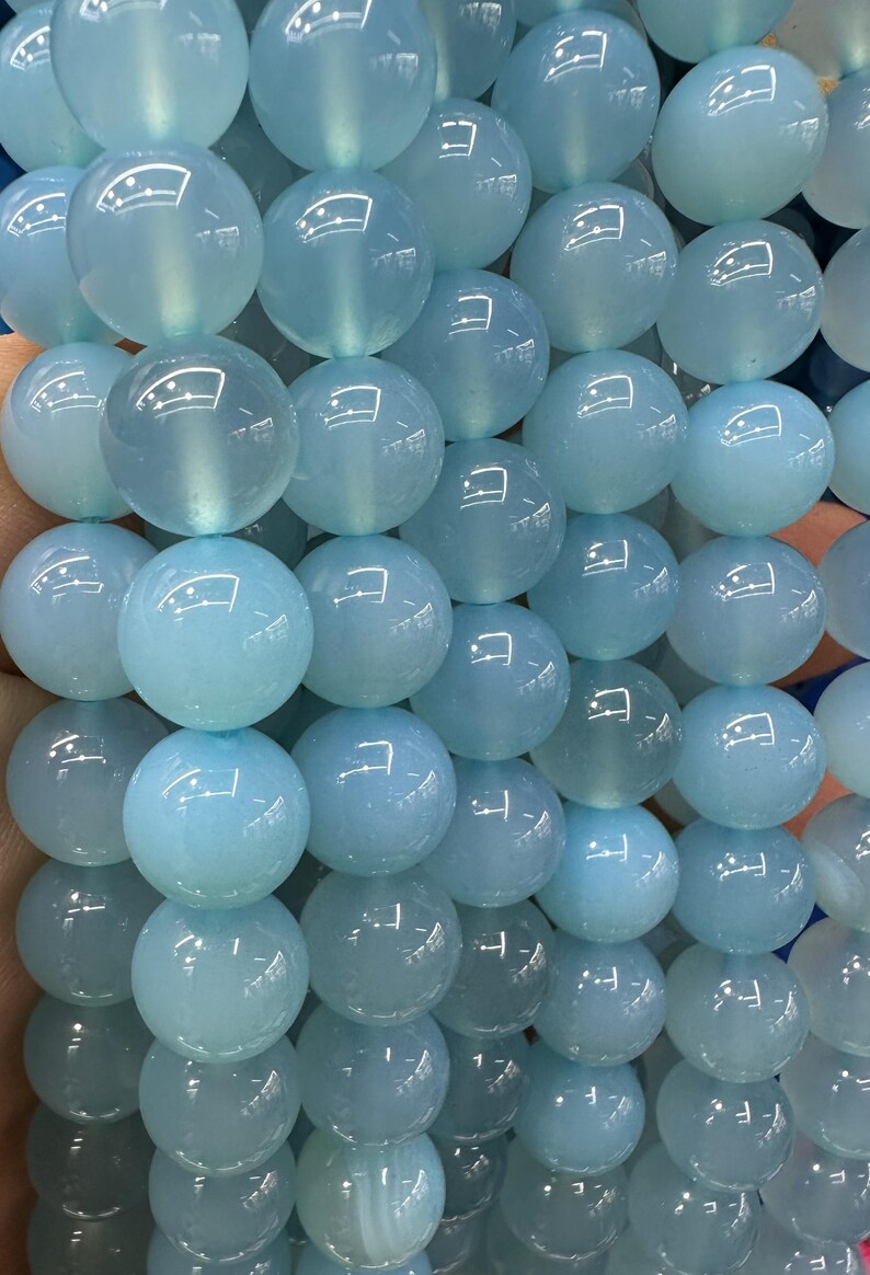 May include: Close-up of a strand of light blue, round gemstone beads. The beads are smooth and translucent, with a subtle sheen. The beads are strung together, creating a full, textured appearance. The beads are approximately 10mm in diameter.