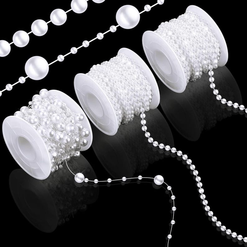 May include: White pearl bead strands and spools against a black background. The strands vary in pearl size, with some on spools and others loose. The pearls have a glossy finish, and the spools are white.