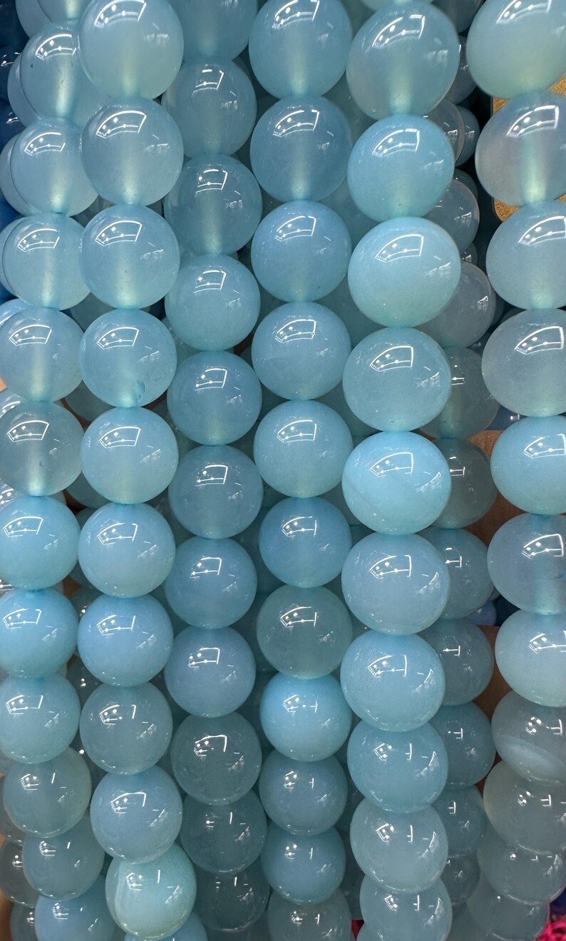 May include: Strands of light blue, round gemstone beads. The beads have a smooth, polished surface and are strung together, creating a shimmering effect. The beads are a soft, translucent color, perfect for jewelry making.