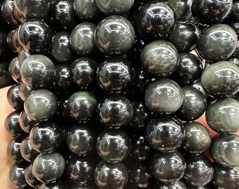 Könnte beinhalten: Nahaufnahme eines Stapels schwarzer Obsidianperlen mit grünem, irisierendem Schimmer. Die Perlen sind rund und glatt und in einem zufälligen Muster angeordnet.
