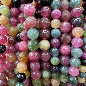 May include: A close-up of strands of colorful gemstone beads. The beads are round and vary in shades of pink, green, yellow, and black. The beads are strung together, creating a textured and vibrant display.