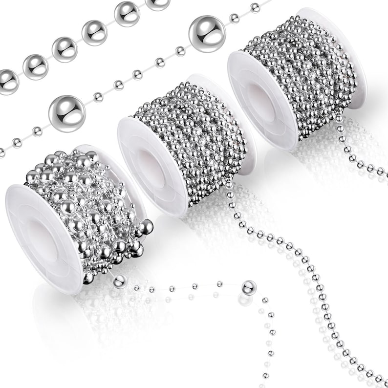 May include: Several spools of silver ball chain are displayed against a white background. The chain is unwound in sections, showing individual beads. The spools are white and the beads are shiny and reflective. The chains are used for jewelry making and crafts.