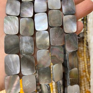 May include: Strand of iridescent, rectangular mother-of-pearl beads in shades of gray, silver, and green. The beads are held in a hand, with other strands of beads visible in the background. The beads are approximately 1 cm square.