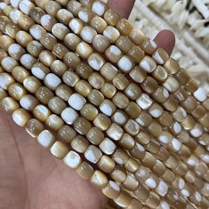 Peut inclure: Gros plan sur des rangées de perles carrées beiges et blanches. Les perles ont un aspect nacré et sont tenues dans une main. Ces perles sont probablement destinées à la fabrication de bijoux.