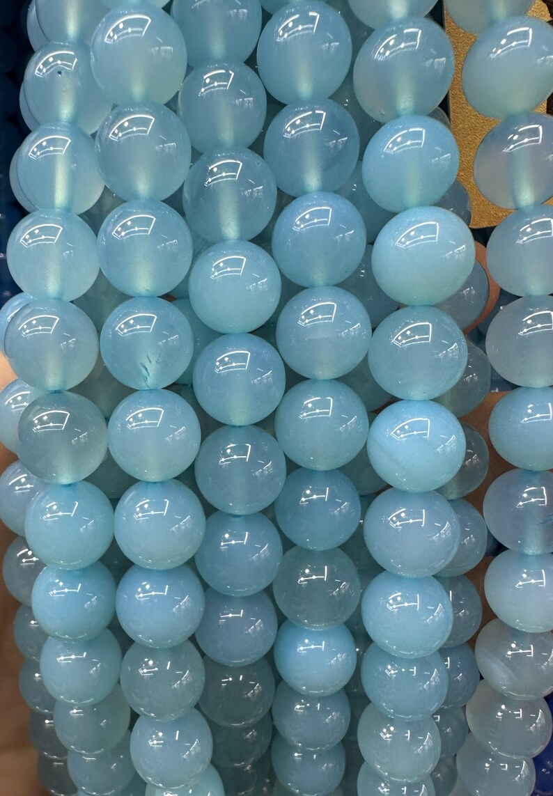 May include: Strand of light blue, translucent gemstone beads. Each round bead has a polished surface, reflecting light. The beads are strung together, creating a necklace or bracelet component. The color is a soft, calming blue.