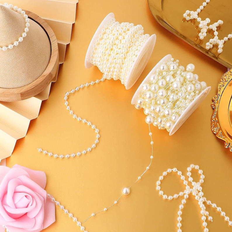 May include: Overhead shot of craft supplies on a gold surface. Two spools of off-white pearl beads are visible, with strands of pearls extending from them. A pink rose and a pearl necklace are also in the frame, suggesting jewelry or craft themes.