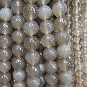 May include: Close-up of strands of grey and white round beads. The beads are smooth and polished, reflecting light. They are strung together, creating a layered effect. These beads are likely used for jewellery making, and are measured in centimetres.