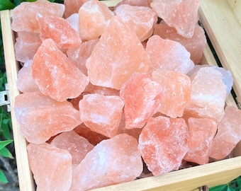 Puede incluir: Una caja de madera llena de numerosos trozos de sal rosa del Himalaya. Los cristales de sal varían en tamaño y forma, mostrando una gama de tonos rosados. La caja es de madera clara y tiene un asa trenzada.