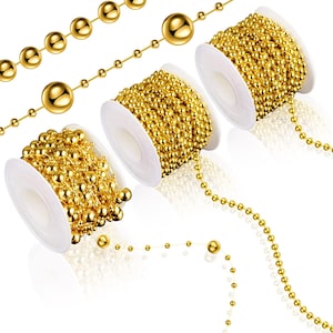 May include: Gold-colored ball chain on white spools and loose strands. The chains feature small, evenly spaced spheres, creating a decorative and shiny effect. The spools are filled with coiled chain, and some strands are unraveled.