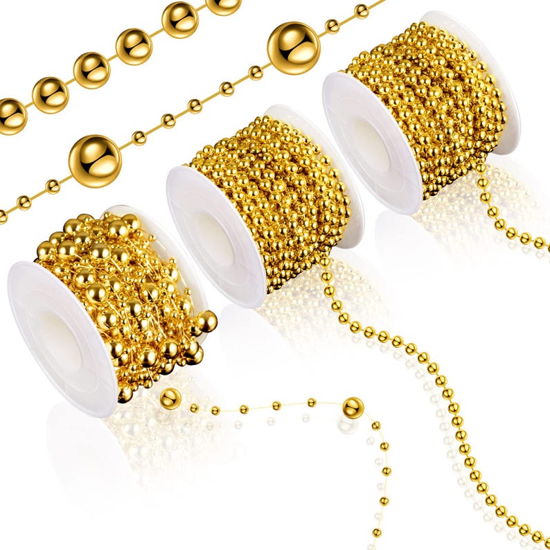 May include: Gold-colored ball chain on white spools and loose strands. The chains feature small, evenly spaced spheres, creating a decorative and shiny effect. The spools are filled with coiled chain, and some strands are unraveled.