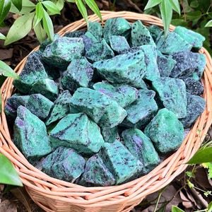 May include: A woven basket overflowing with raw ruby zoisite stones. The stones are a mix of green and black with pink flecks. The basket is surrounded by green leaves and rests on a natural surface.