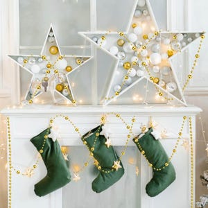 May include: A festive Christmas mantelpiece decorated with two white star-shaped ornaments filled with silver, gold, and white baubles. Three green stockings with star accents hang below, adorned with gold bead garlands.