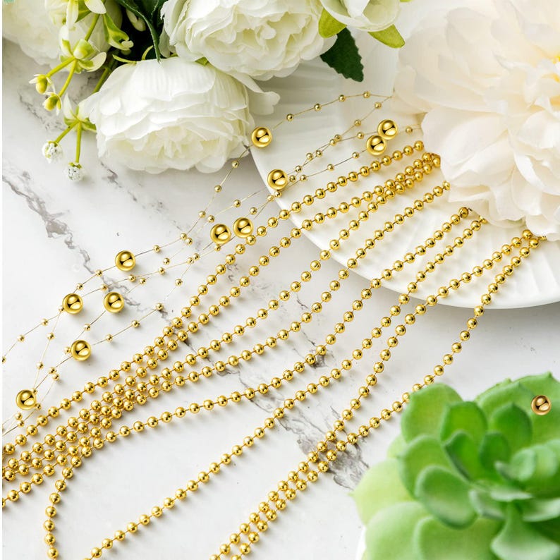 May include: Gold beaded garland strands with varying bead sizes. The strands are arranged on a white plate and a marble surface, with white flowers and greenery in the background. The garland is suitable for decorative purposes.