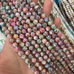 May include: Strands of round beads in a variety of colors, including pink, green, and blue. The beads have a speckled pattern and are held in a hand. Other strands of beads are visible in the background.