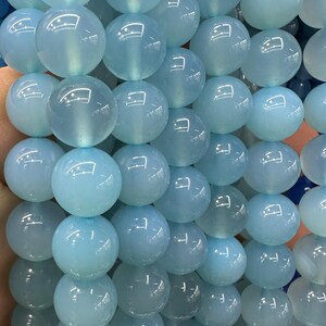 May include: Close-up of a strand of light blue, translucent, round beads. The beads have a glossy finish and are strung together, creating a textured surface. The beads are approximately 10mm in diameter, suitable for jewelry making.