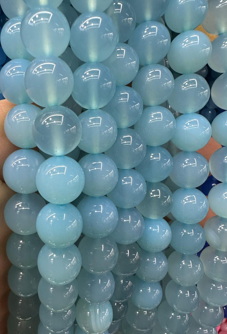 May include: Close-up of a strand of light blue, translucent, round beads. The beads have a glossy finish and are strung together, creating a textured surface. The beads are approximately 10mm in diameter, suitable for jewelry making.