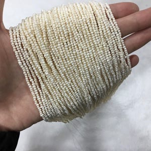 Puede incluir: Colección de pequeñas cuentas redondas de color blanquecino, probablemente perlas, ensartadas en múltiples hebras. Las cuentas están muy juntas, creando una apariencia densa y texturizada. Las hebras se mantienen unidas, lo que sugiere que son para la fabricación de joyas.