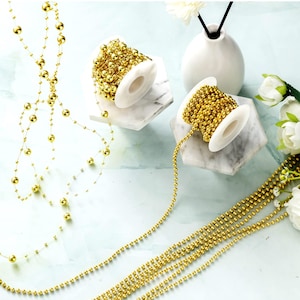 May include: Gold beaded garland and strands of beads are displayed. Two spools of gold beads sit on white and gray marble-like stands. The garland is draped and arranged, showcasing the decorative potential of the beads.