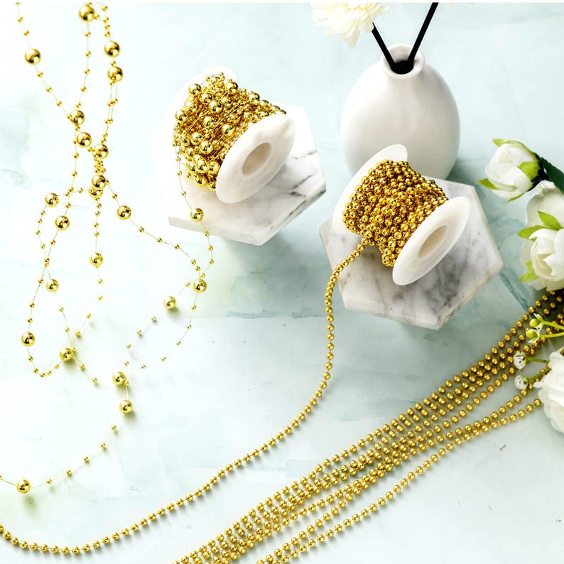 May include: Gold beaded garland and strands of beads are displayed. Two spools of gold beads sit on white and gray marble-like stands. The garland is draped and arranged, showcasing the decorative potential of the beads.