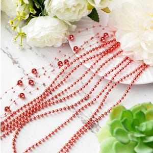 May include: Red beaded garland strands with larger accent beads. The garland is arranged on a white plate with white flowers and a green succulent in the background. Decorative beads for crafts and decor.