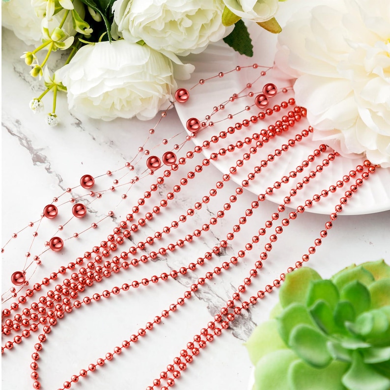 May include: Red beaded garland strands with larger accent beads. The garland is arranged on a white plate with white flowers and a green succulent in the background. Decorative beads for crafts and decor.