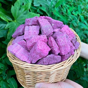 Puede incluir: Una cesta de mimbre llena de golosinas rectangulares de color rosa brillante. La cesta se sostiene frente a un fondo de follaje verde. Las golosinas parecen ser un refrigerio o un alimento.