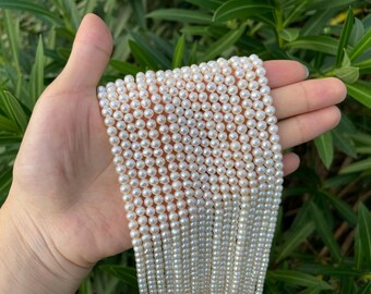 Könnte beinhalten: Mehrere Stränge weißer Perlen. Die Perlen sind klein, rund und haben einen glänzenden Glanz. Die Stränge werden in einer Hand gehalten, mit grünem Laub im Hintergrund. Die Perlen sind ein klassisches Schmuckstück.