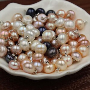 May include: A collection of freshwater pearl beads in various colors, including white, peach, and dark gray. Each bead has a silver-colored metal loop attached. The pearls are displayed in a white, shallow dish.